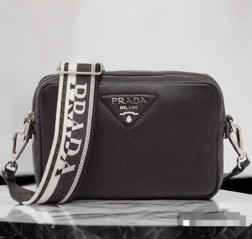 Bolso crossbody Prada Milano en cuero graneado color beige/taupe y versión camel, con correa ancha de tela bordada con el logotipo de la marca, placa triangular metálica frontal y herrajes plateados.