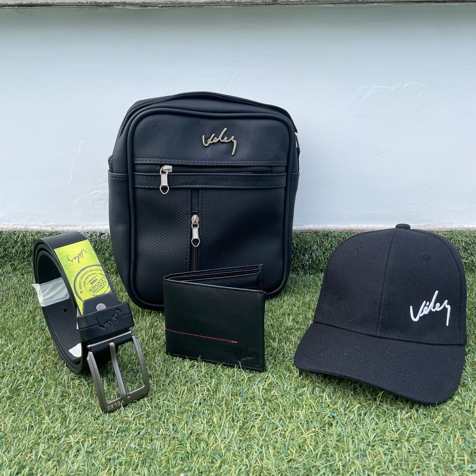 Set de accesorios ejecutivos que incluye un bolso manos libres con textura microperforada, una billetera de cuero liso con costura diagonal, un cinturón con hebilla metálica y una gorra deportiva con marca metalica