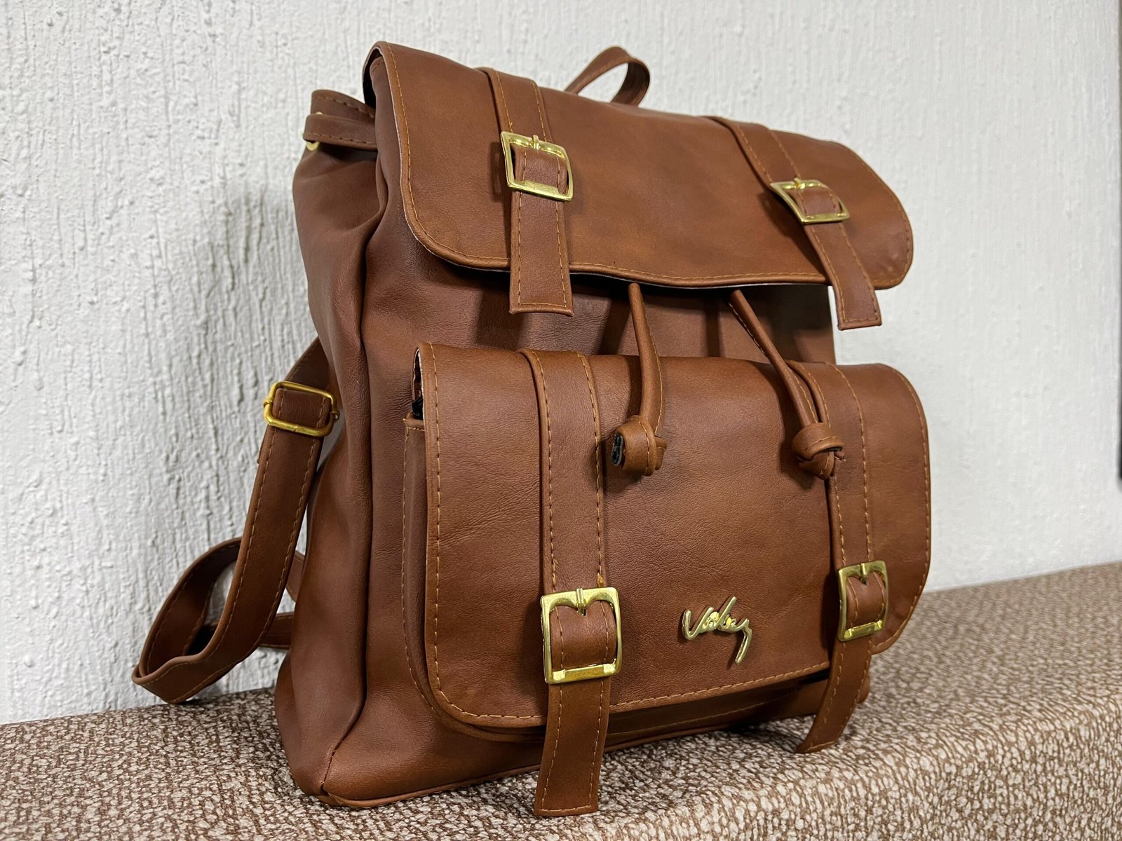 Mochila de silueta estructurada con doble solapa, correas decorativas con hebillas metálicas doradas, cierre de cordón ajustable y herraje de firma en relieve.
