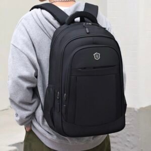 Mochila ejecutiva negra de diseño minimalista con panel frontal liso, escudo metálico plateado en la parte superior, bolsillos laterales de malla y correas acolchadas