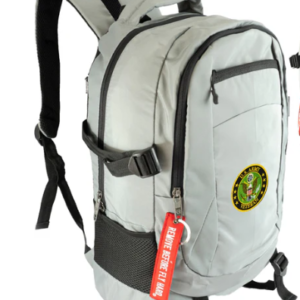 Morral gris claro de estilo aviador con parche circular de "U.S. Army Veteran", etiqueta roja colgante "Remove Before Fly Hard", bolsillo lateral de malla y correas de compresión.
