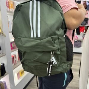 Morral Adidas de color verde oliva con tres rayas verticales blancas en la parte superior, logotipo de Adidas en el bolsillo frontal, bolsillos laterales de malla negra y un llavero decorativo de la marca.