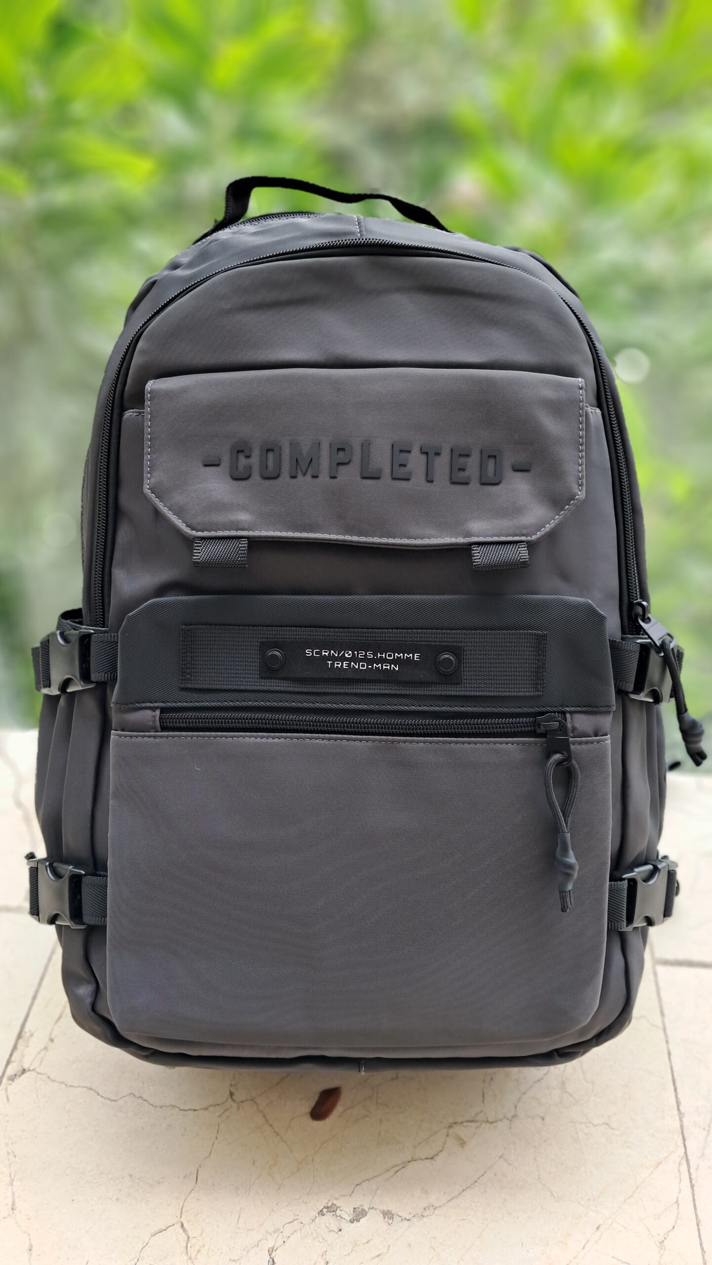 Morral técnico gris oscuro con relieve "COMPLETED" en el bolsillo frontal, correas de compresión laterales, múltiples compartimentos con cremallera y asa superior reforzada.