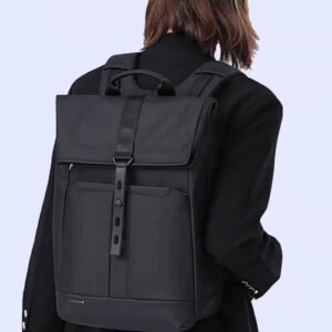 Morral negro de cuero sintético mate con diseño de solapa superior, dos correas verticales con hebillas doradas, cordón de ajuste interno y correas de hombro delgadas, sobre un suelo de mosaico artístico.