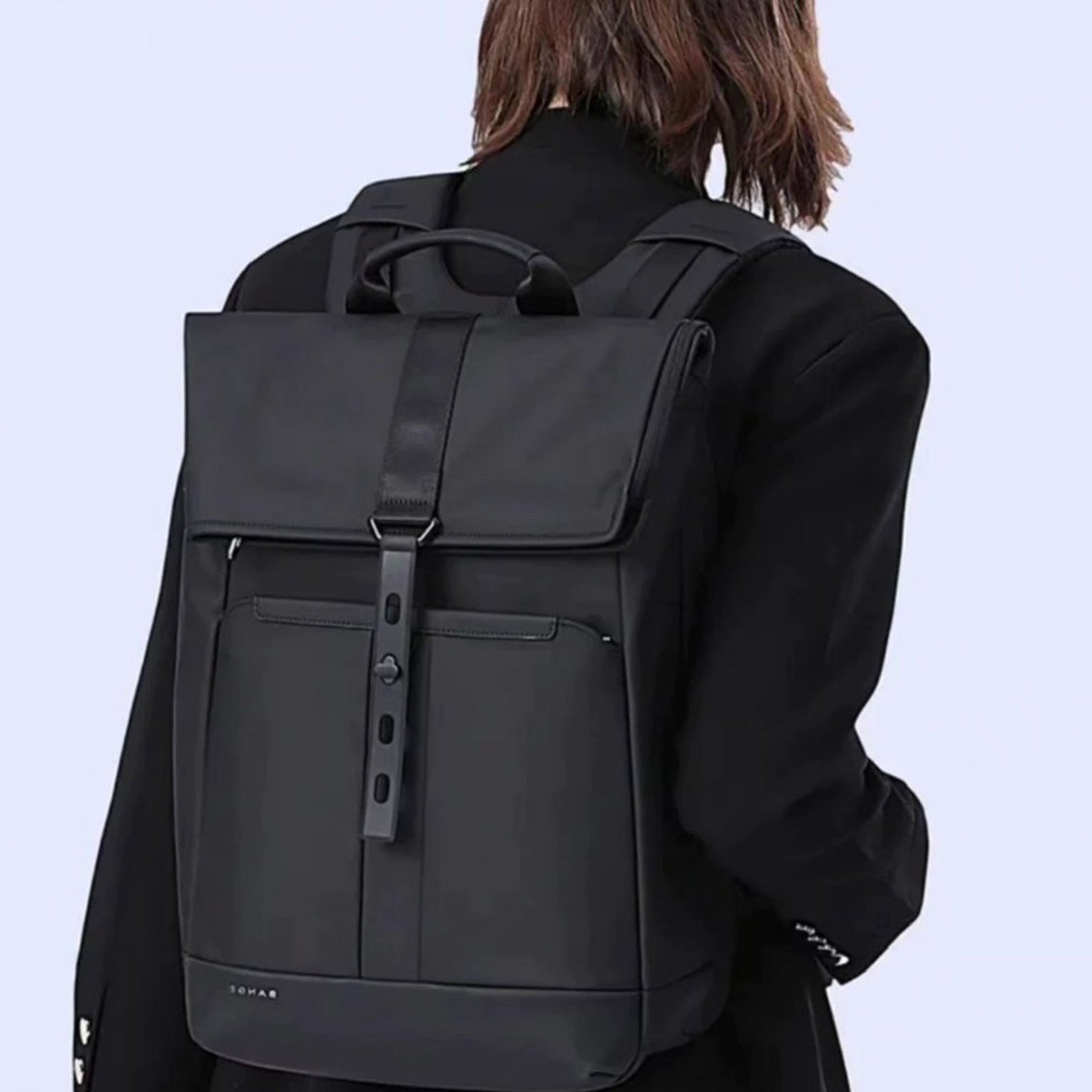 Morral negro de cuero sintético mate con diseño de solapa superior, dos correas verticales con hebillas doradas, cordón de ajuste interno y correas de hombro delgadas, sobre un suelo de mosaico artístico.